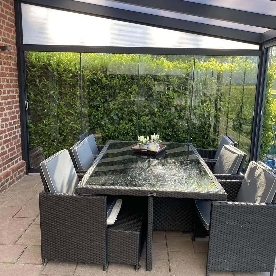 Tuintafel onder een veranda met glazen schuifwanden en een groene haag